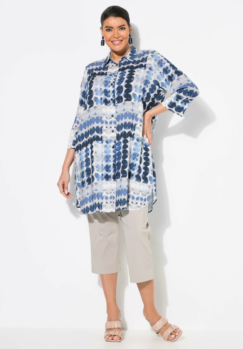 Blaue und weiße Bluse mit geometrischen Mustern, Knopfleiste vorne und 3/4-Ärmeln, kombiniert mit beigen Cropped-Hosen und hellbeigen Absatzsandalen.