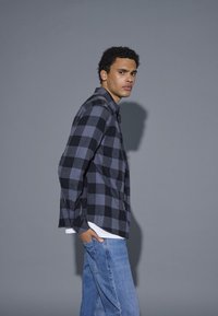 Camicia a quadri nera e grigia con chiusura a bottoni, abbinata a jeans di denim blu chiaro. Tessuto morbido, vestibilità rilassata e maniche lunghe.
