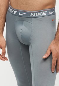 Άνδρας που φορά γκρι κολάν Nike με επώνυμη ζώνη και μικρό πορτοκαλί σήμα Nike στο μηρό, στέκεται με τα χέρια χαλαρά στο πλάι.