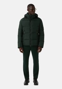 Padded green jacket with a hood, vertical quilted design, zippered pockets, paired with green pants and black shoes. Minimalist style.