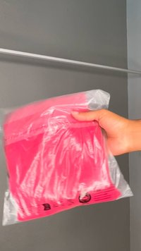 Jaskro różowy element odzieży zapakowany w przezroczystą plastikową torbę, trzymany w dłoni. Torba ma wydrukowane symbole i tekst na dole.
