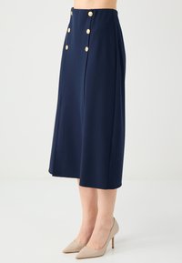 Gonna midi blu navy con un design plissettato, dotata di bottoni dorati in vita e tessuto a trama liscia. Indossata con scarpe con tacco alto color nude.