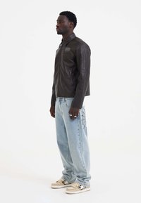 Black leather jacket with a pointed collar, zippered pockets, paired with light blue denim jeans featuring a spiderweb design. Cream sneakers.