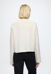Pull court couleur crème avec manches longues, confectionné en matière douce, doté d'un col rond et d'une coupe décontractée. Pantalon noir visible.