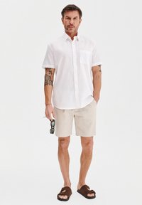 Biała krótka koszula z kieszonką, połączona z jasnobeżowymi szortami w paski oraz brązowymi sandałami. Model trzyma okulary przeciwsłoneczne w lewej ręce.