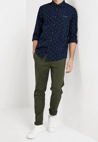 Marineblaue Hemd mit Knöpfen, bedeckt mit weißen und roten Tupfen; grüne, eng geschnittene Hose; weiße Sneakers; hochgekrempelte Ärmel; kleine Brusttasche.