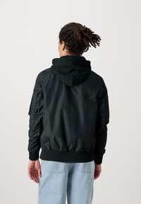 Černá bomber bunda s kapucí, prošívaná textura, žebrované manžety a lemování. Disponuje hladkým povrchem a přiléhavým střihem. Ukažte zadní pohled.