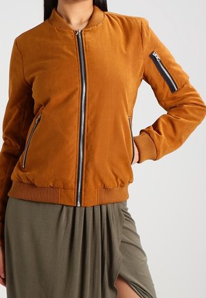 Femme portant une veste bomber couleur rouille avec des poches zippées et des poignets côtelés, associée à une jupe verte olive avec une fente à l'avant.