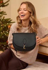Borsa a tracolla in pelle nera con una patta curva, hardware dorato e una forma strutturata, tenuta da una persona che indossa un maglione beige.