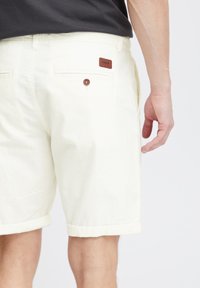 Shorts albe din in cu mansete răsucite, având un buzunar posterior și o etichetă din piele maro. Țesătura are o finisare netedă și texturată.