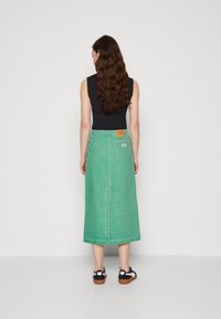 Grön denim midi-kjol med rak passform, vertikal sömnad och ett lapptyg på baksidan. Tillsammans med svarta sneakers, sedd bakifrån.