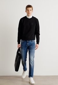 Black sweatshirt with a small chest pocket, paired with blue jeans and white sneakers. The model holds a black tote bag.