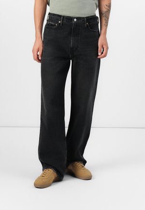 Person iført løstsiddende sorte jeans og beige sneakers, der står mod en ensfarvet grå baggrund med afslappede hænder ned langs siderne.