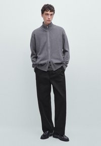 Maglione zip grigio chiaro con colletto e polsini a costine, abbinato a pantaloni neri larghi e scarpe nere su uno sfondo neutro.
