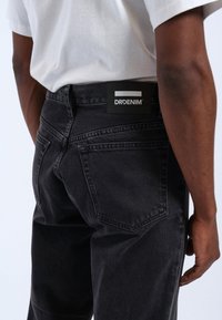 Jeans de mezclilla negra con corte recto, con bolsillos traseros y una etiqueta en la cinturilla. Llevados con una camiseta blanca, mostrando la vista trasera.