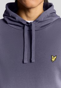Lila hoodie med snörning och en liten gul broderad logotyp på bröstet. Tillverkad av ett mjukt tyg med en slät yta.