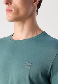 T-shirt in cotone verde teal con scollo rotondo, caratterizzato da un piccolo logo in rilievo quadrato "BOSS" nella parte sinistra del petto.