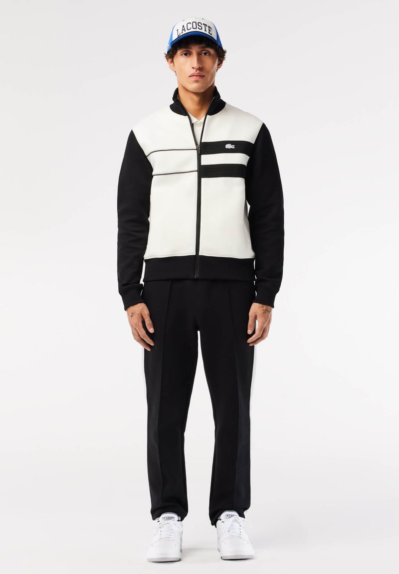 Veste zippée pour homme en blanc et noir, avec un design texturé et des rayures horizontales. Un pantalon noir et des baskets blanches complètent l'ensemble.