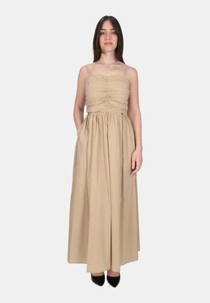 Donna in piedi di fronte indossa un abito lungo smanicato beige arricciato con tasche e tacchi neri a punta su uno sfondo semplice.