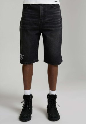 Nogi w czarnych dżinsowych szortach do kolan z białym haftem przy brzegu, białe skarpety sportowe oraz czarne sznurowane buty na szarym tle.