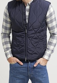 Navy quilted vest with a high collar, zip closure, and front pockets, paired with a light plaid shirt and blue jeans.