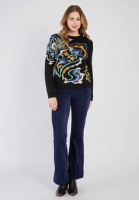 Pull noir avec un motif floral coloré, associé à un pantalon en velours côtelé bleu foncé évasé et des bottines noires.