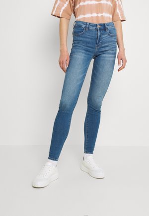 Personne portant un jean skinny bleu, des baskets blanches et une chemise à manches courtes rayée marron et blanche, debout devant un fond clair uni.