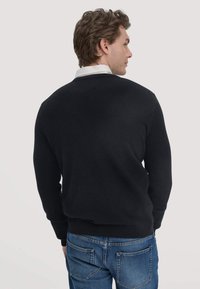 Schwarzer Pullover mit Rundhalsausschnitt, gerippten Bündchen und Saum. Über einem weißen Hemd mit Kragen getragen, kombiniert mit blauen Jeans. Weiche Textur, lässiges Design.