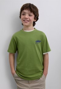 Vihreä lyhythihainen t-paita, jossa on valkoinen kaulus ja sininen kuorma-autoaihe. Pehmeää puuvillamateriaalia ja rento istuvuus, yhdistetty beigen väristen shortseihin.