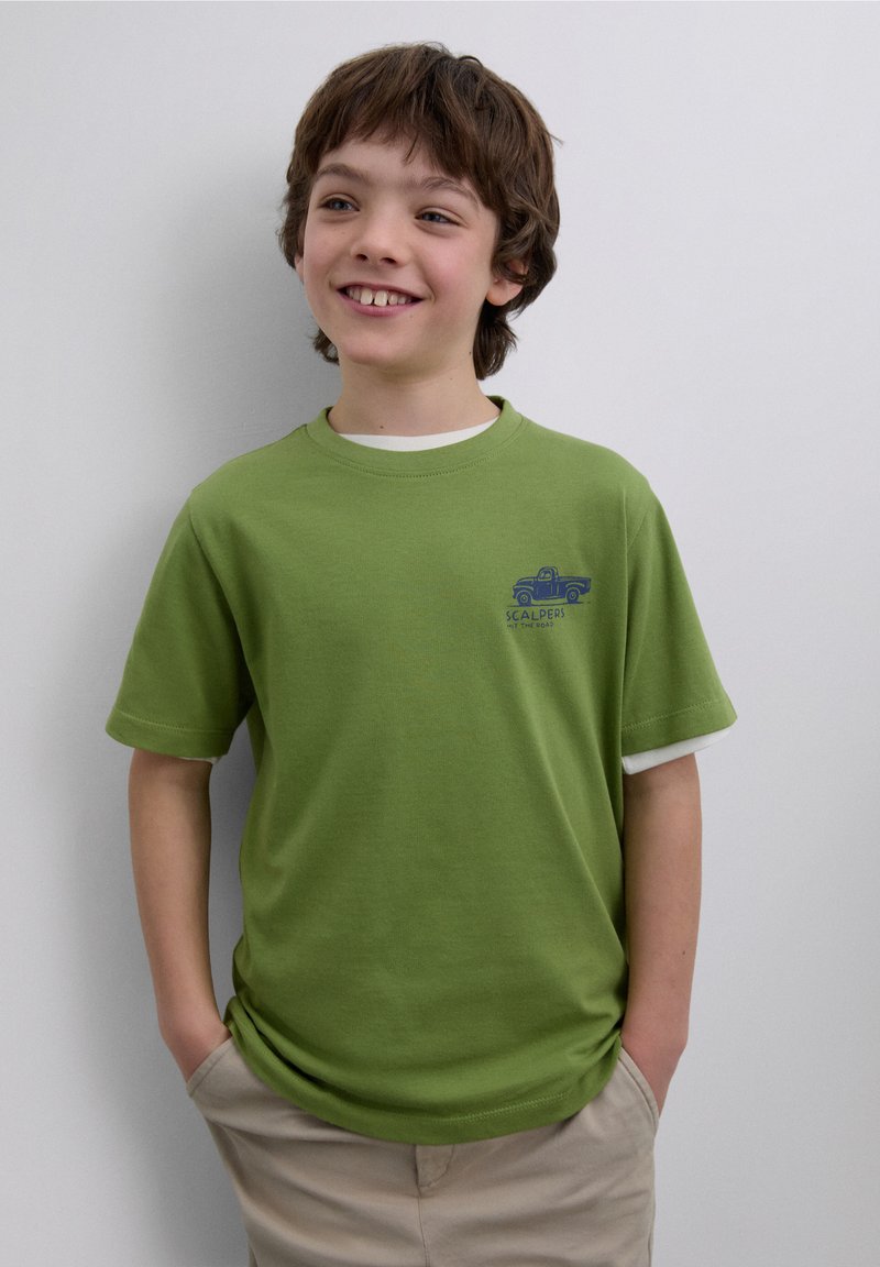 Vihreä lyhythihainen t-paita, jossa on valkoinen kaulus ja sininen kuorma-autoaihe. Pehmeää puuvillamateriaalia ja rento istuvuus, yhdistetty beigen väristen shortseihin.