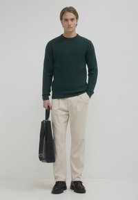 Dunkelgrüner strukturierter Strickpullover, cremefarbene weit geschnittene Hose, schwarze Leder Schuhe und eine schwarze Schultertasche mit einem langen Tragegurt.