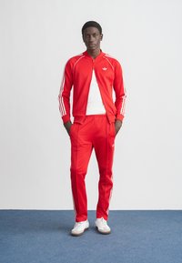 Röd träningsdräkt med jacka med dragkedja och matchande byxor med vita randiga detaljer på ärmarna och sidorna, kombinerad med vita sneakers.