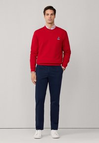 Felpa rossa con collo rotondo e logo frontale, abbinata a pantaloni blu scuro e sneaker bianche. Texture liscia e design semplice.