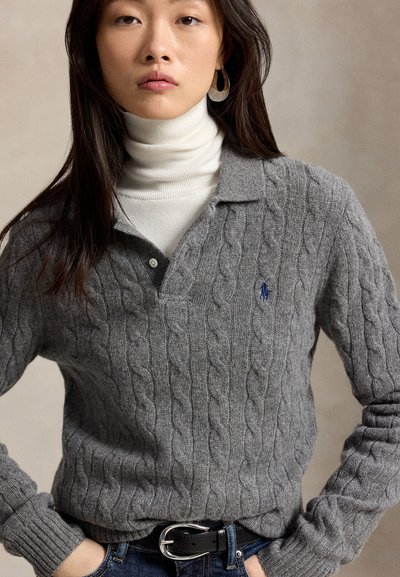 Jeune femme aux longs cheveux foncés porte un col roulé blanc sous un pull en maille torsadée gris avec un col et un logo sur la poitrine.