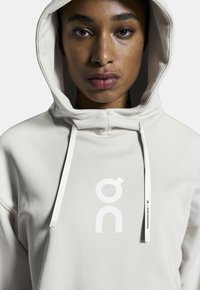 Hoodie beige clair avec une texture douce, arborant un grand logo blanc et des cordons de serrage. Le design comprend une poche kangourou sur le devant et une capuche.