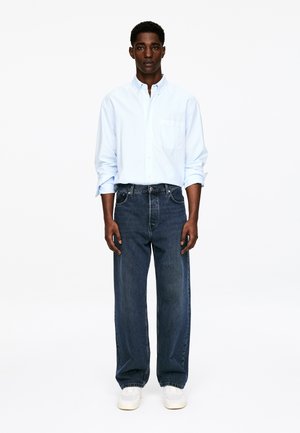 Homme debout portant une chemise bleu clair à boutons avec les manches retroussées, un jean large noir, et des baskets blanches sur fond blanc.