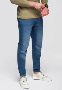 Joggers de mezclilla azul tapered con puños elásticos, que presentan un lavado desgastado y sutiles detalles de desgaste. Combinados con zapatillas blancas.