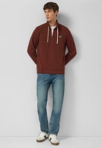 Pullover bordeaux con mezza zip e strato interno bianco, con cordini crema e logo sul petto, abbinato a jeans azzurri e sneaker bianche.