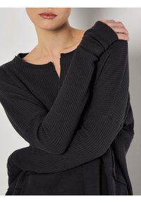 Top lungo nera in maglia waffle con scollatura ampia e bordi grezzi. Presenta maniche aderenti e una silhouette rilassata.