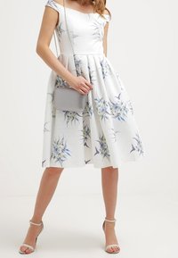 Robe blanche à épaules dénudées avec imprimés floraux en bleu et vert. Jupe longue, tissu texturé, associée à un sac à main gris et des sandales à lanières.