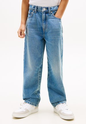 Kind draagt losse lichtblauwe spijkerbroek en witte sneakers, met één hand in de zak en de andere ontspannen langs het lichaam.