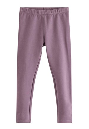 Barn med krøllet hår iført mørkebrun hættetrøje, hvid prikket skjorte, lilla leggings, lyserøde sokker og beige sko står mod hvid baggrund.