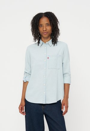 Kvinde med krøllet hår iført en lyseblå skjorte med knapper og op rullede ærmer samt mørkeblå jeans, der står foran en ensfarvet baggrund.