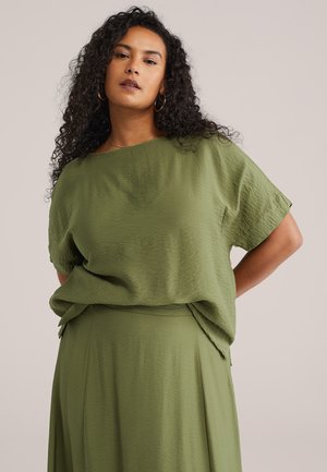 Kvinde med krøllet mørkt hår iført olivengrøn, tekstureret løs top og matchende nederdel mod en ensfarvet baggrund.