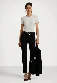 Femeie îmbrăcată cu un tricou alb mulat, pantaloni negri slim, curea neagră, sandale cu toc negre, ținând în mână un sacou negru cu logo brodat.