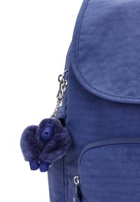 Blue fabric backpack with a flap closure, featuring a furry gorilla keychain and silver hardware. Textured surface with zipper pockets.