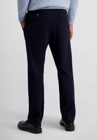 Man in donkerblauwe broek met achterzakken en zwarte schoenen, staande tegen een effen lichte achtergrond.