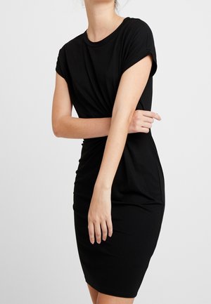 Femme en robe noire ajustée à manches courtes avec des détails froncés, debout devant un fond clair uni, les bras croisés devant elle.