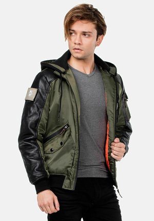 Olivgrüne Bomberjacke mit schwarzen Lederärmeln, Kapuze und Reißverschlüssen; hat ein oranges Innenfutter und einen Aufnäher am linken Arm.