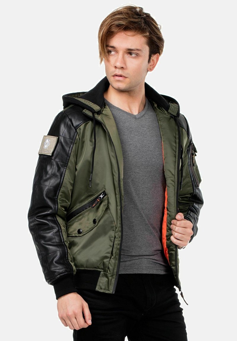 Olivgrüne Bomberjacke mit schwarzen Lederärmeln, Kapuze und Reißverschlüssen; hat ein oranges Innenfutter und einen Aufnäher am linken Arm.